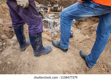 two workers and a foreman are standing next to the building site, the ground, excavated trench, pit, the feet of builders and workers in dirty boots and a shabby work robe
