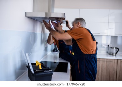 Two workers, handyman in uniform installing or repairing a kitchen extractor, replacing filter in cooker hood. Construction, maintenance and repair concept. Side view. Horizontal shot