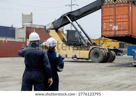 Two workers in hard hats oversee the loading of shipping containers at an industrial port using heavy machinery.