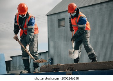 Two Workers In Hard Hats, Work Clothes And A Medical Mask Work With Shovels At A Construction Site. Hard Physical Labor. An Authentic Scene At Work