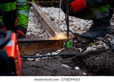 Two workers in high visibility gear weld a corroded steel rail on crushed stone as sparks fly. Boots, a rebar stake with green tape, and power cables sit in muddy soil in daytime.