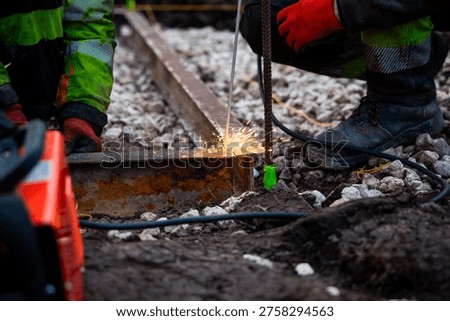 Two workers in high visibility gear weld a corroded steel rail on crushed stone as sparks fly. Boots, a rebar stake with green tape, and power cables sit in muddy soil in daytime.