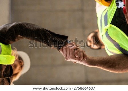Two workers hold hands in a gesture of agreement at a construction site during daylight hours. Both are wearing safety gear and showing teamwork in their work environment.