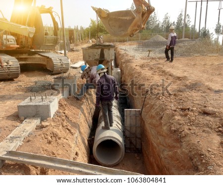 Two workers are installing concrete drains on the side of the road, which is a large power plant construction. Backhoe lifting of concrete pipe drainage system.