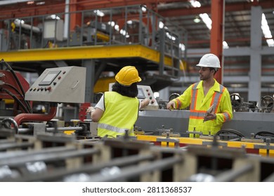 Two workers interact while standing near machines in a manufacturing facility. One worker gestures while holding a clipboard. The other listens attentively.