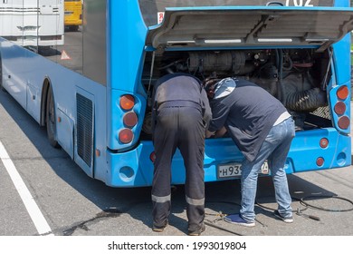Two workers are leaning forward and under the raised hood are repairing the engine of a modern blue passenger bus. Scattered tools and spare parts can be seen on the asphalt nearby.