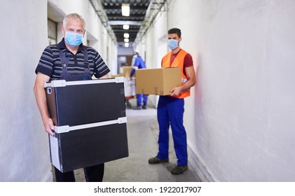 Two Workers With Masks Because Of Covid-19 Carry Packages Into The Warehouse Of A Factory