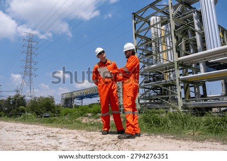 Two workers in orange jumpsuits and helmets stand near industrial equipment. They look at a document and discuss tasks. Power lines run in the background on a sunny day.