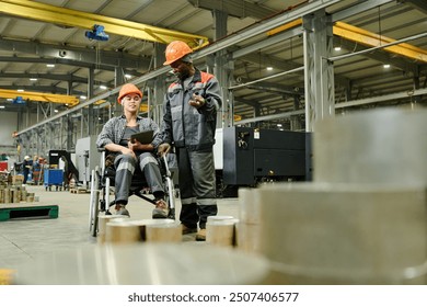 Two workers in protective gear collaborating in factory environment, one person seated in wheelchair holding clipboard and device, surrounding machinery and metal components visible - Powered by Shutterstock