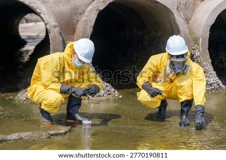 Two workers in protective gear crouch by a stream collecting water samples. They wear yellow suits, gloves, and masks. The setting shows large industrial pipes and water around them.
