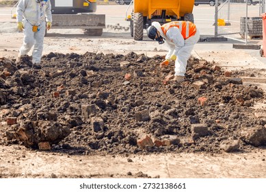 Two workers in protective gear remove asbestos from contaminated ground filled with bricks at construction site
