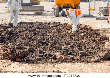 Two workers in protective gear remove asbestos from contaminated ground filled with bricks at construction site