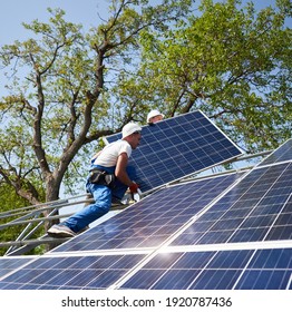 Two workers in protective helmet on tall steel platform mounting heavy solar photo voltaic panel on green tree and blue sky background. Exterior solar panel system installation, dangerous job concept. - Shutterstock ID 1920787436