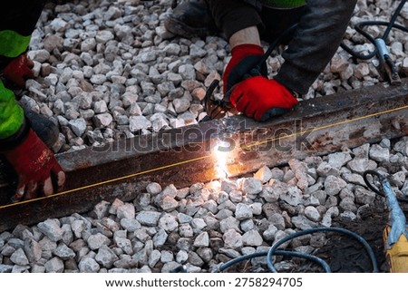 Two workers in red gloves weld a weathered steel rail on crushed stone ballast as sparks flare and a cutting torch follows a taut guide line during daylight track work.
