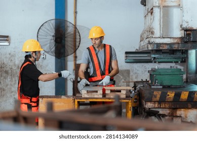 Two workers in safety gear are using equipment in a factory. One worker points at a machine while the other prepares materials on a table.