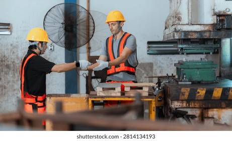 Two workers in safety gear are using equipment in a factory. One worker points at a machine while the other prepares materials on a table.