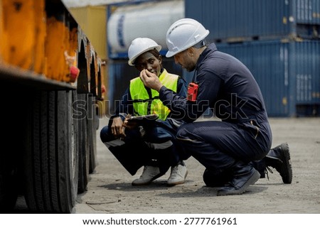 Two workers in safety helmets and vests inspect a large vehicle tire at an industrial shipping yard with stacked containers in the background.