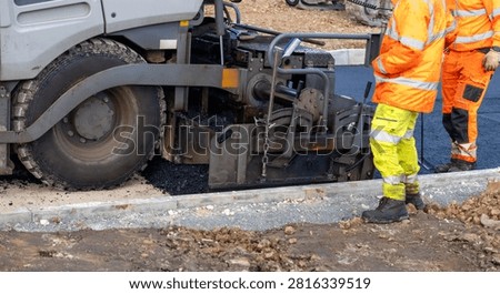 Two workers stand beside a paver machine that is laying asphalt on a road. Work is taking place in a construction area during the day.