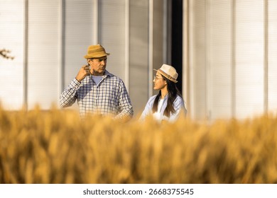 Two workers stand in a wheat-covered area in front of large silos, actively discussing upcoming tasks.