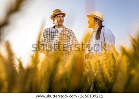Two workers stand in a wheat-covered area in front of large silos, actively discussing upcoming tasks.