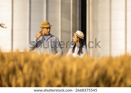 Two workers stand in a wheat-covered area in front of large silos, actively discussing upcoming tasks.