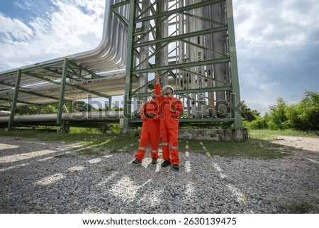 Two workers supervisor dressed in orange safety suits and white helmets observing pipeline project at construction site. engineer at industrial tube oil petrochemical.