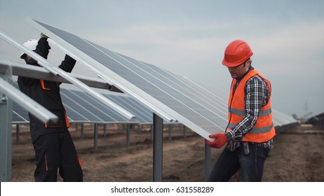 Two Workers In A Uniform And Hardhat Install Photovoltaic Panels On A Metal Basis On A Solar Farm