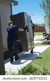 Two workers using shoulder dolly lifting straps, gloves delivering new refrigerator in modern home via concrete walkway in Princeton, Texas. Appliance delivery service truck parked upfront street. USA