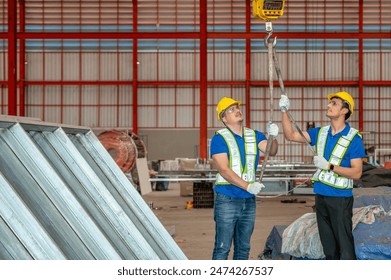 Two workers wearing hard hats and safety vests use a chain hoist to lift a heavy load in a warehouse - Powered by Shutterstock