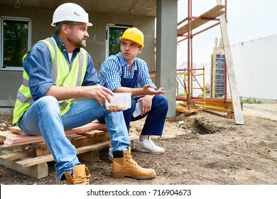 Two Workers Wearing Protective Helmets Taking Break From Work And Enjoying Lunch While Sitting Outdoors, Unfinished Building On Background