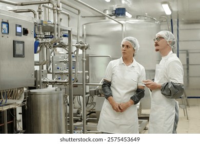 Two workers wearing safety nets and white lab coats observing machinery and equipment in food manufacturing facility. They seem to be discussing operations or examining equipment