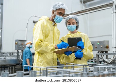 Two workers in yellow protective suits examine data together. One holds a clipboard, the other uses a tablet. Industrial facility features pipes and machinery
