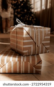 Two wrapped gift boxes are stacked on a wooden floor with a blurred Christmas tree in the background.