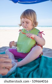 Two Years Old Child With Green Bib Sitting On Towel Down Parasol Umbrella At Beach Eating Tuna Salad With Orange Plastic Fork From Tupperware