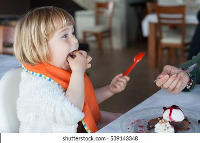 Two Years Old Child With Orange Plastic Spoon Eating With Hand In Mouth Sharing With Woman A Piece Of Chocolate Cake With Vanilla Ice Cream At Restaurant