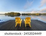 Two yellow Adirondack chairs on a wooden dock greet the tranquil Muskoka summer morning, facing the blue waters of a lake. Across the way, cottages nestled among green trees complete the idyllic scene