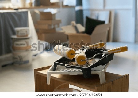 Two yellow paintrollers on stack of plastic trays standing on wooden piece of furniture in front of camera in living room
