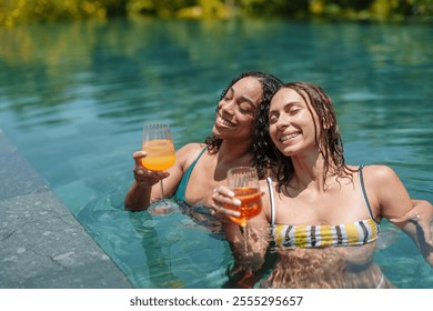 Two young adult women, one of diverse ethnicity and the other Caucasian, smile brightly while sipping beverages in a sunny pool setting, their attire casual swimwear. - Powered by Shutterstock