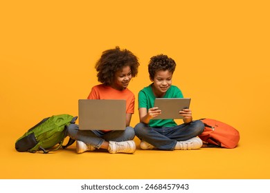 Two young African American children absorbed in a laptop and tablet. Their backpacks rest beside them, hinting at a break from school activities or a moment of leisure after classes. - Powered by Shutterstock