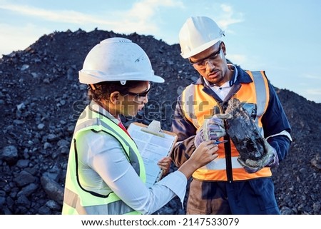 Two young African mine workers wearing protective wear are discussing coal quality while on site at a coal mine