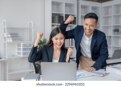 Two young Asian business people successful excited raised hands rejoicing with a laptop computer in a modern office. - Powered by Shutterstock