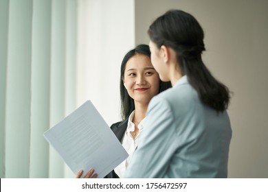 Two Young Asian Business Women Standing By Window Having A Discussion In Office