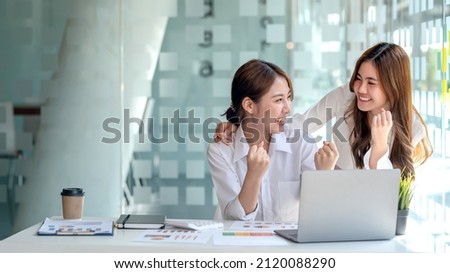 Two young Asian businesswomen show joyful expression of success at work smiling happily with a laptop computer in a modern office.