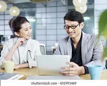 Two Young Asian Corporate People Discussing Business In Office Using Tablet Computer.