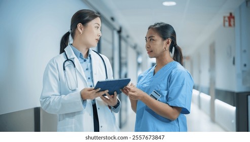 Two Young Asian Female Medical Professionals Standing in a Bright Hospital Corridor with Other People, Engaged in a Serious Discussion While the Doctor Reviews Patient Details on a Tablet Computer - Powered by Shutterstock