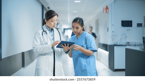 Two Young Asian Female Medical Professionals Standing in a Bright Hospital Corridor with Other People, Engaged in a Serious Discussion While the Doctor Reviews Patient Details on a Tablet Computer - Powered by Shutterstock