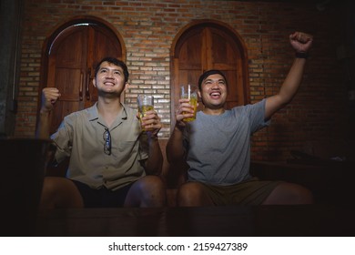 Two Young Asian Friends Happily And Happily Drinking Beer In Front Of The TV Cheering For Football. Friends Clink Beer At Home At Night To Cheer On Football.