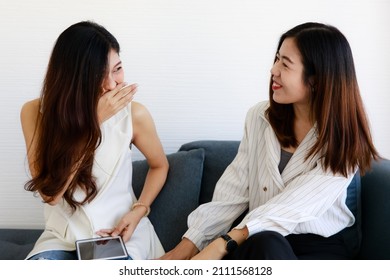 Two young Asian girls wearing office business style clothes sitting together on sofa, talking funny stories, smiling and laughing with relaxing action. Female colleagues pose in secret gossip style.