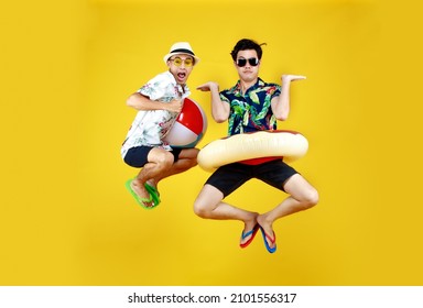 Two young Asian guys in summer casual with hat and sunglasses jumping up high with funny gesture with beach ball and ring float as excited and happy to having fun playing at swimming pool on vacation.