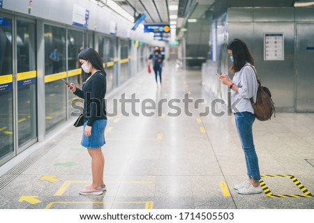 Two young asian woman wearing protective face mask stand in line with social distancing during waiting train in subway due to Coronavirus or COVID-19 outbreak situation in all of landmass in the world